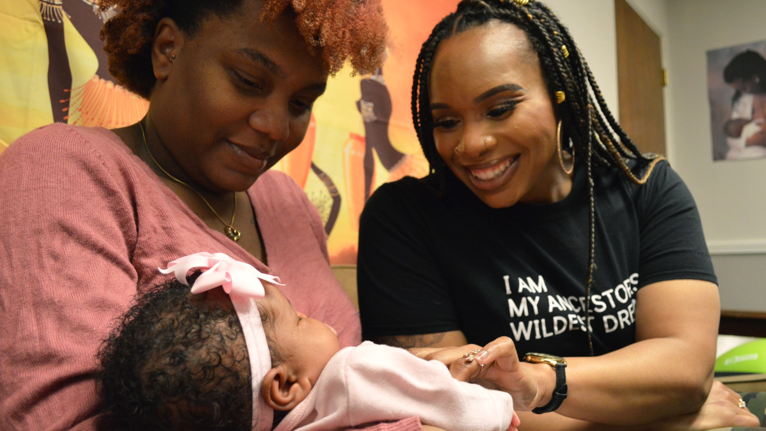 A woman in a pink shirt holds a baby as another woman looks on