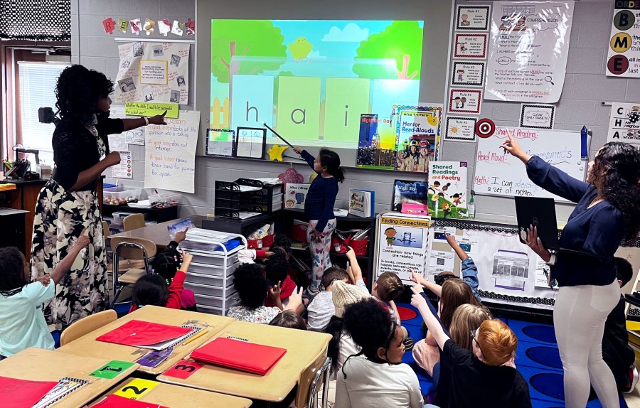 A young girl holds a stick and points out large letters spelling hair. two teachers teach behind her