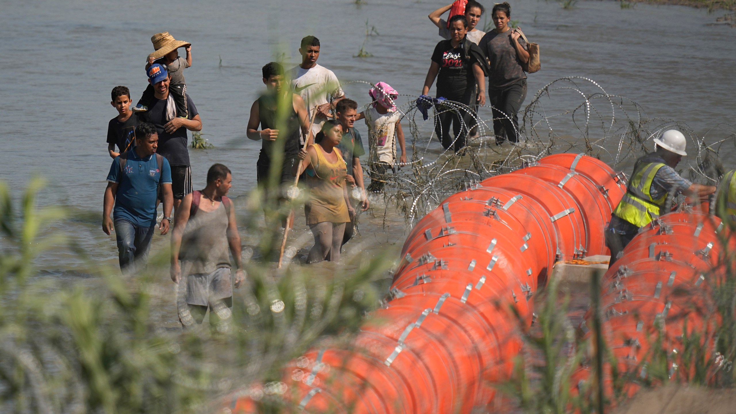 Migrants trying to enter the U.S. from Mexico approach the site where workers are assembling large buoys to be used as a border barrier along the banks of the Rio Grande in Eagle Pass, Texas, Tuesday, July 11, 2023. The floating barrier is being deployed in an effort to block migrants from entering Texas from Mexico. (AP Photo/Eric Gay)