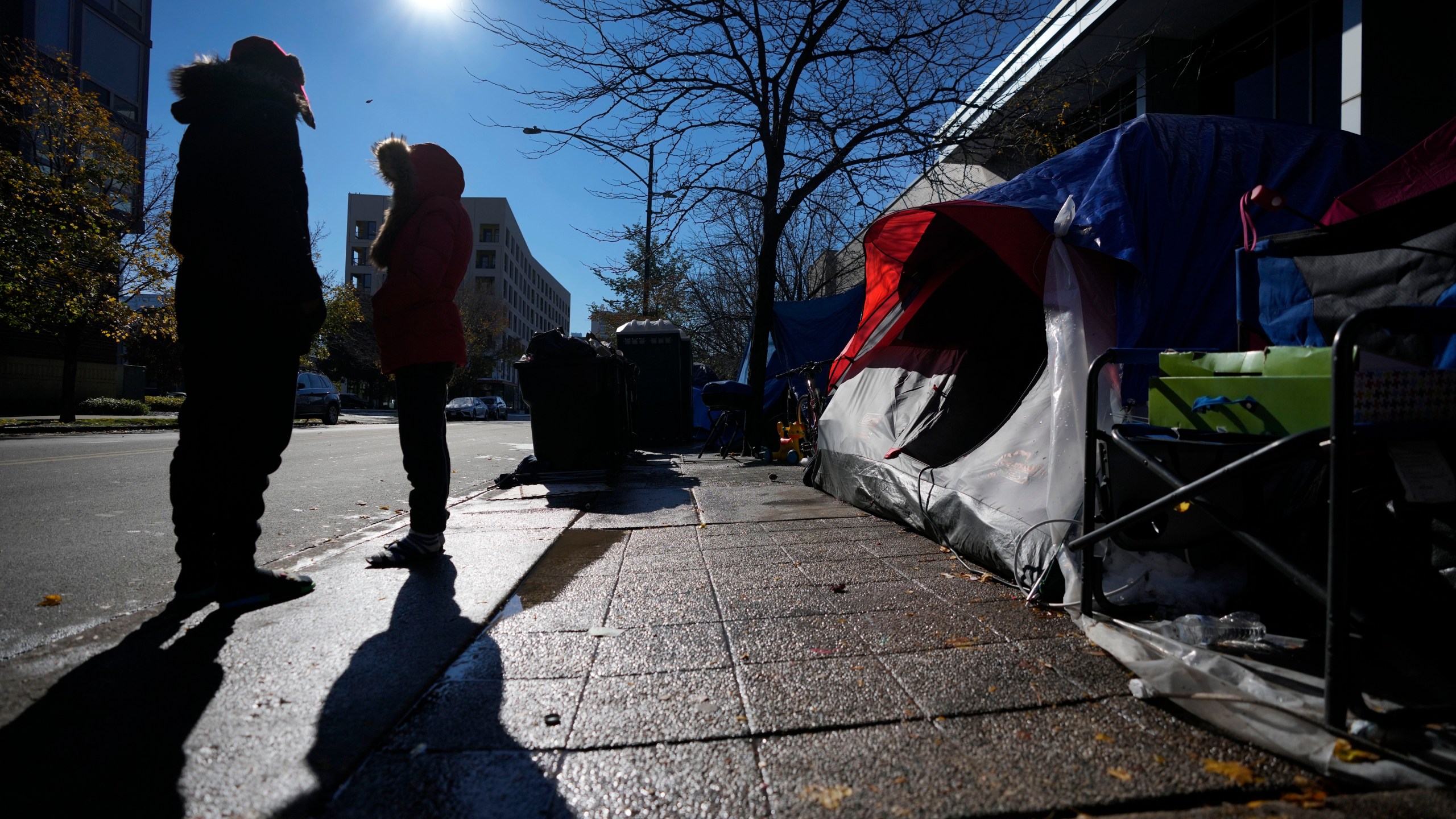 A man and woman stand in the late morning sun near their tent in a small migrant community Wednesday, Nov. 1, 2023, near a Northside police station in Chicago. (AP Photo/Charles Rex Arbogast)