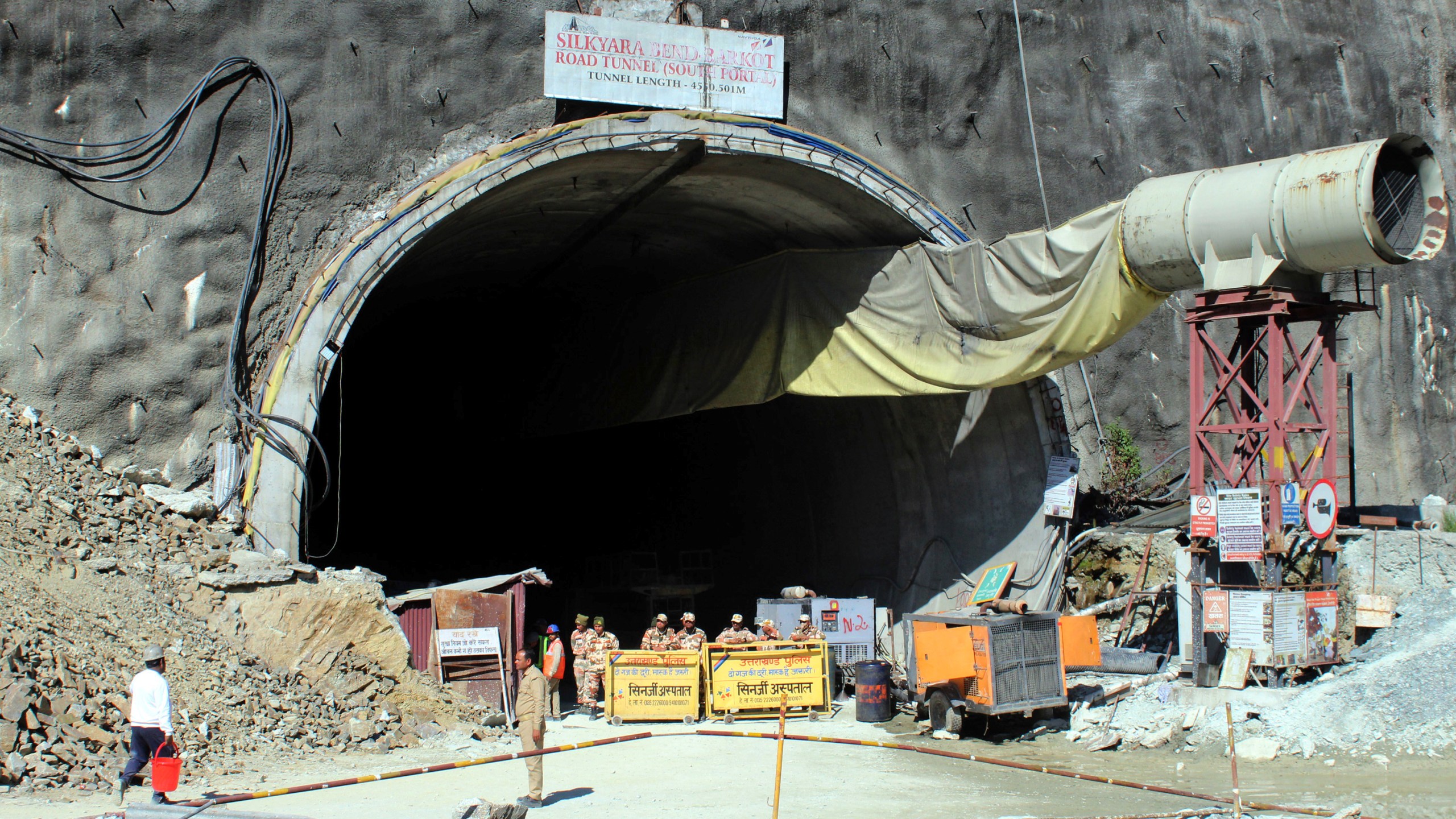 Security personnel stand guard near the entrance to the site of an under-construction road tunnel that collapsed in mountainous Uttarakhand state, India, Wednesday, Nov. 15, 2023. Rescuers have been trying to drill wide pipes through excavated rubble to create a passage to free 40 construction workers trapped since Sunday. A landslide Sunday caused a portion of the 4.5-kilometer (2.7-mile) tunnel to collapse about 200 meters (500 feet) from the entrance. It is a hilly tract of land, prone to landslide and subsidence. (AP Photo)