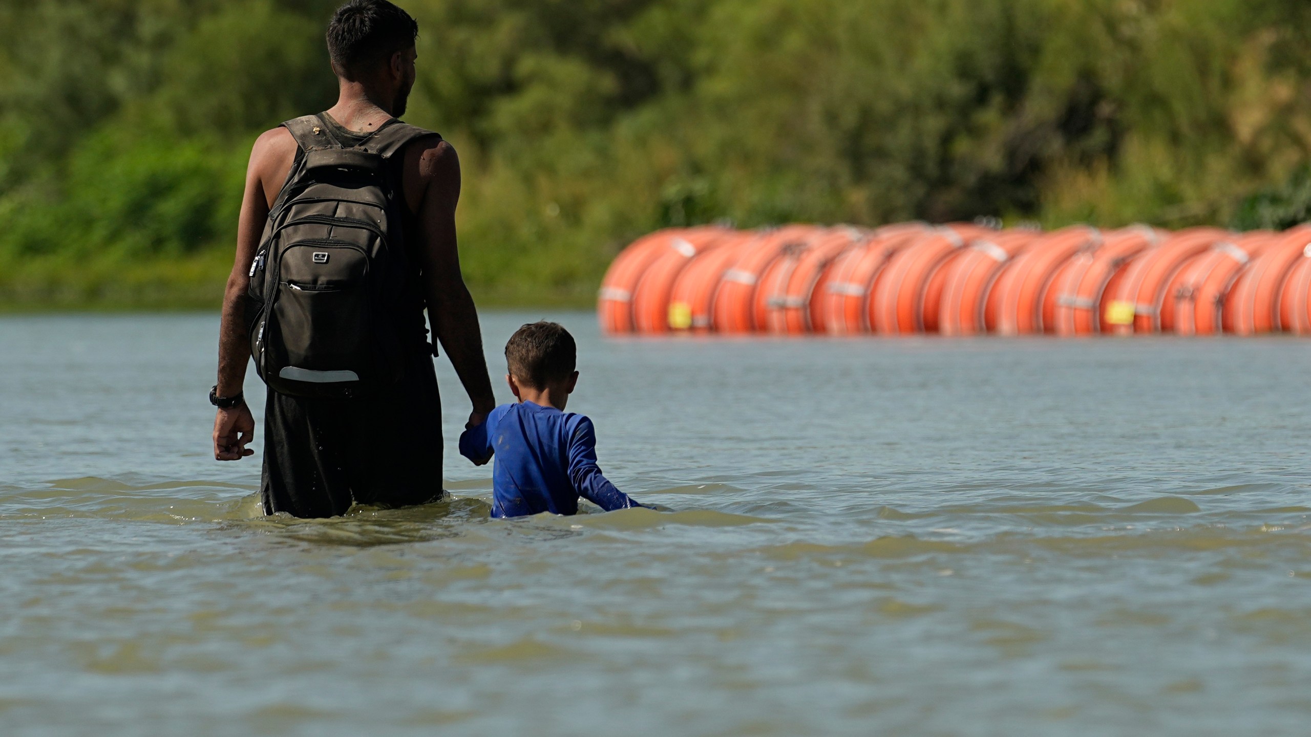 FILE - Migrants walk past large buoys being used as a floating border barrier on the Rio Grande, Aug. 1, 2023, in Eagle Pass, Texas. Texas must move a floating barrier on the Rio Grande that drew backlash from Mexico, a federal appeals court ruled Friday, Dec. 1, 2023, dealing a blow to one of Republican Gov. Greg Abbott's aggressive measures aimed at stopping migrants from entering the U.S. illegally. (AP Photo/Eric Gay, file)