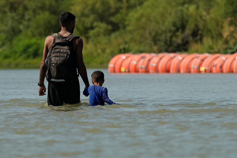 FILE - Migrants walk past large buoys being used as a floating border barrier on the Rio Grande, Aug. 1, 2023, in Eagle Pass, Texas. Texas must move a floating barrier on the Rio Grande that drew backlash from Mexico, a federal appeals court ruled Friday, Dec. 1, 2023, dealing a blow to one of Republican Gov. Greg Abbott's aggressive measures aimed at stopping migrants from entering the U.S. illegally. (AP Photo/Eric Gay, file)