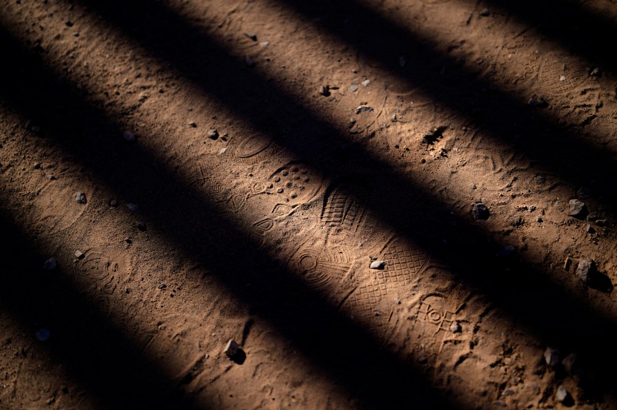 FILE - Footsteps are seen along a road shadowed by the steel columns of the border wall separating Arizona and Mexico, Friday, Dec. 15, 2023, near Lukeville, Ariz. (AP Photo/Gregory Bull, File)