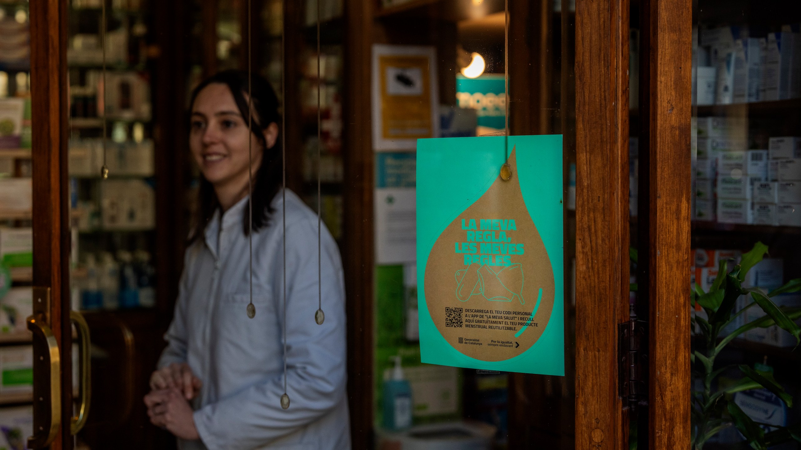 A pharmacist waits for customers next to windows where an advertisement for the new campaign of the Catalan government where you can read "my period, my rules" has been placed, inside a pharmacy in Barcelona, Spain, Tuesday, March 5, 2024. Spain's Catalonia region rolled out this week a pioneering women's health initiative that offers millions of women reusable menstruation products for free. Some 2.5 million women in northeast Spain can receive one menstrual cup, one pair of underwear for periods, and two packages of cloth pads at local pharmacies free of charge. (AP Photo/Emilio Morenatti)