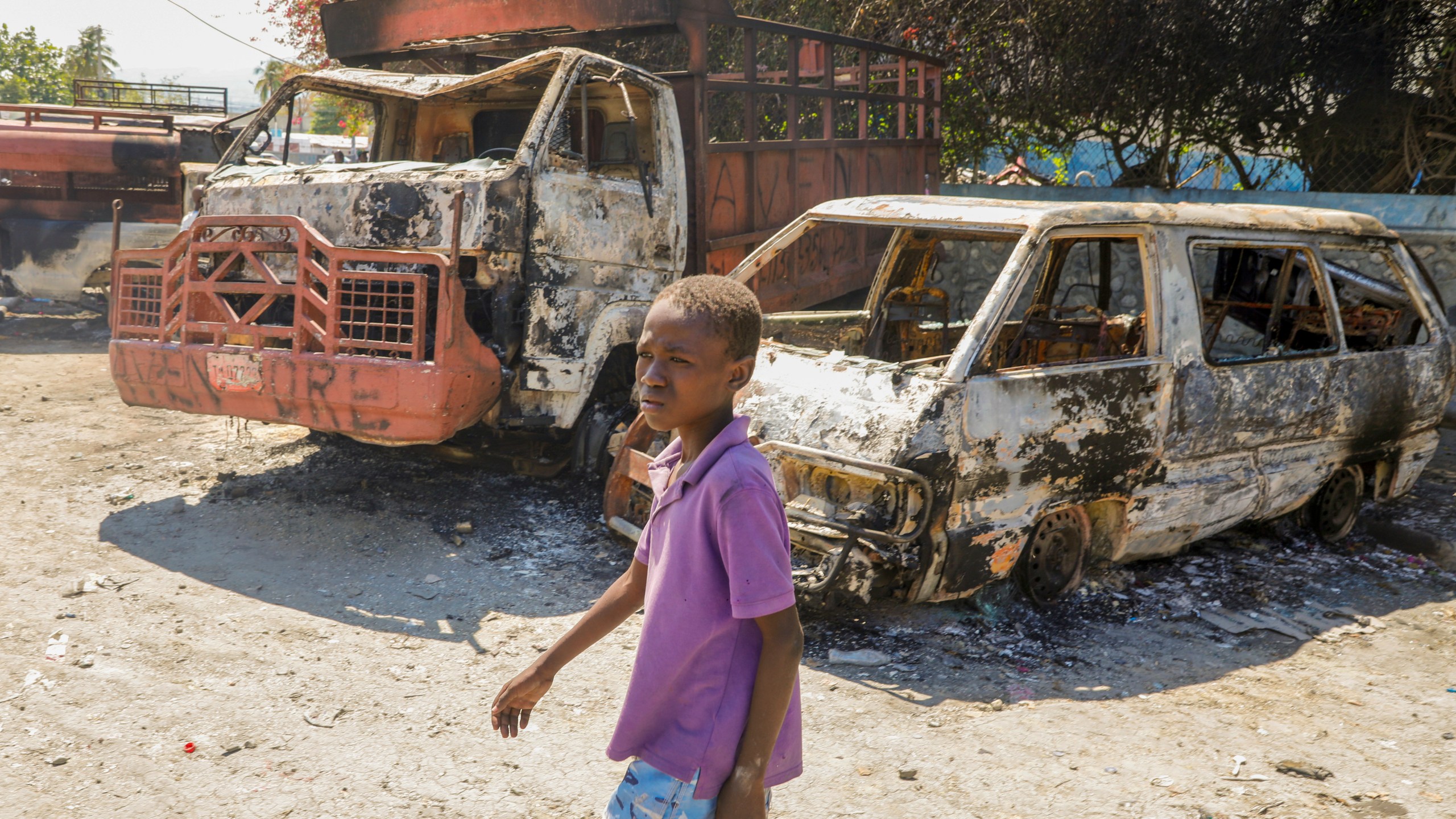 A youth walks by charred cars outside a police station set on fire by armed gangs in Port-au-Prince, Haiti, Tuesday, March 5, 2024. Prime Minister Ariel Henry has been absent since the country's latest and most serious outbreak of violence started the previous week, and armed groups have seized on the power void. (AP Photo/Odelyn Joseph)