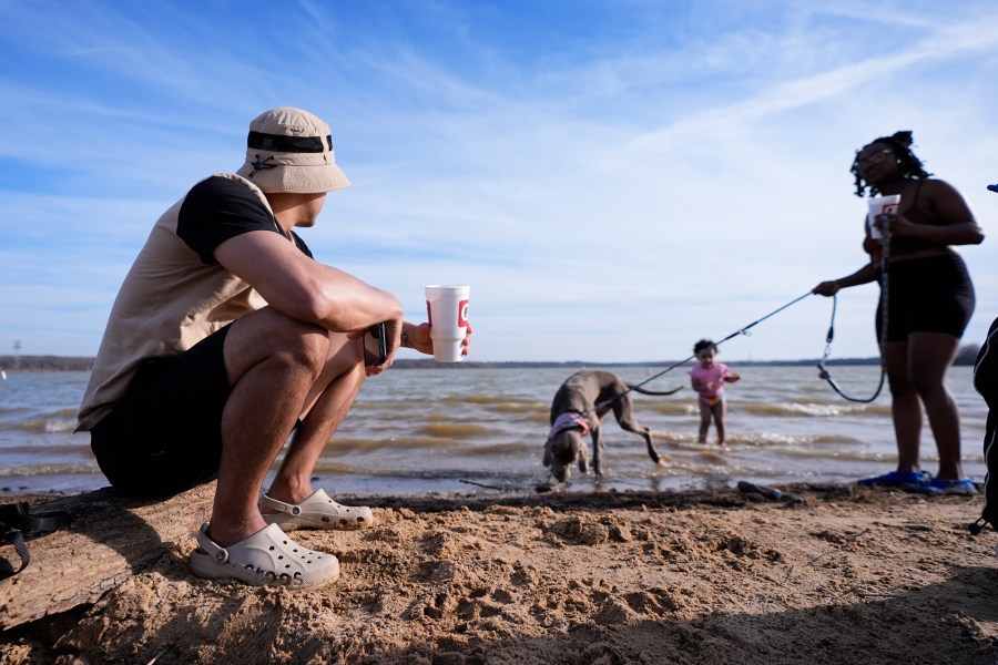 FILE - Victor Marcano, left, sits on a log as he watches his daughter Zhamira Marcano, 3, stand in the water on the shore of Joe Pool Lake with Joanna Clarkley, right, and her dog, Pluto, during an unseasonably warm winter day, Monday, Feb. 26, 2024, in Grand Prairie, Texas. Federal meteorologists on Friday, March 8, have made it official: It's the warmest U.S. winter on record by far. (AP Photo/Julio Cortez, File)