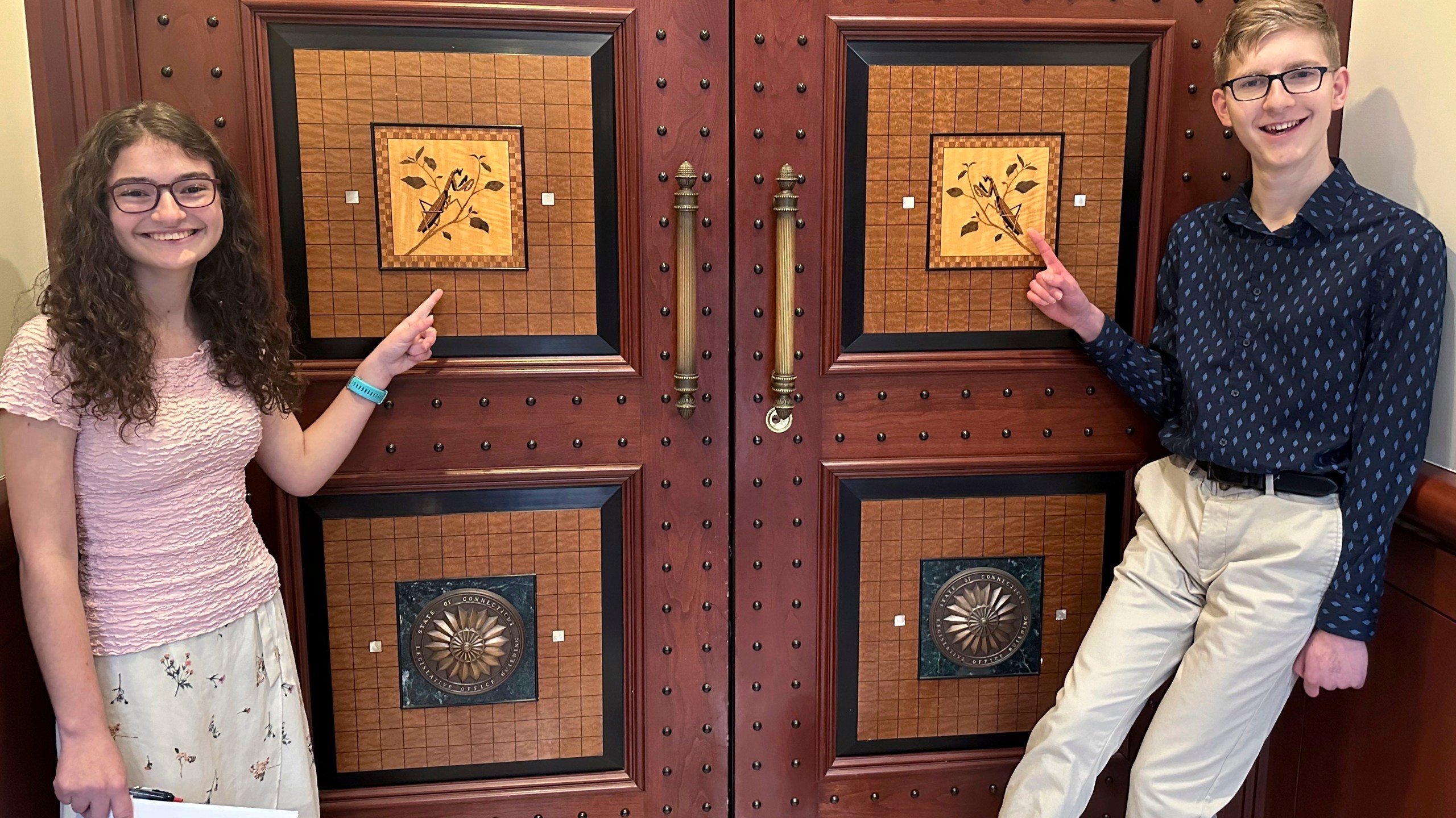 Sue Katherine Boye Williams, 16, a junior at the Watkinson School in Hartford, Conn. and Chris Tracy, 19, a senior at the school, stand near an inlaid design of Connecticut's current State Insect, the European "Praying" Mantis, on the door to a hearing room at Connecticut's Legislative Office Building in Hartford on March 8, 2024. Both testified before a legislative committee, urging lawmakers to replace the non-native praying mantis with the native Spring Azure Butterfly. (AP Photo/Susan Haigh)