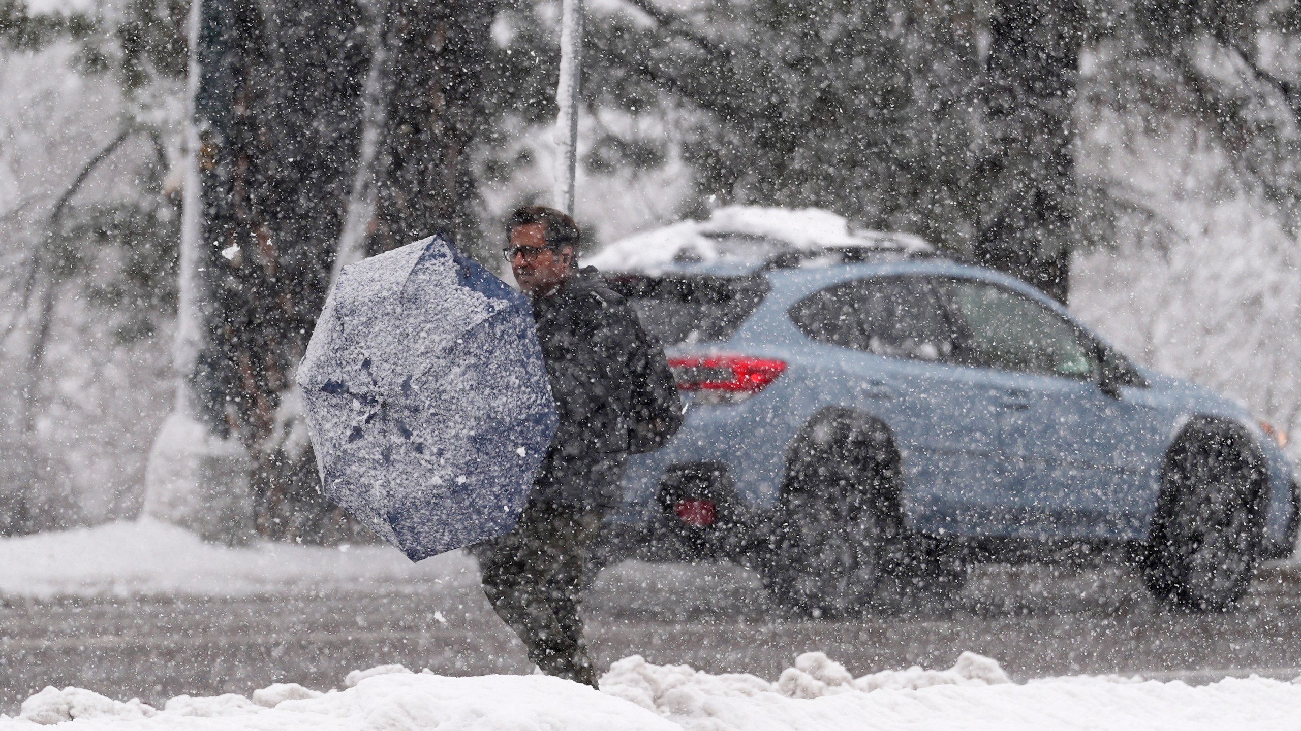 A pedestrian cleans off his umbrella while waiting for a bus at a stop along eastbound Speer Boulevard during a snow stormon Thursday, March 14, 2024, in Denver. Forecasters predict that the storm will persist until early Friday, snarling traffic along Colorado's Front Range communities. (AP Photo/David Zalubowski)