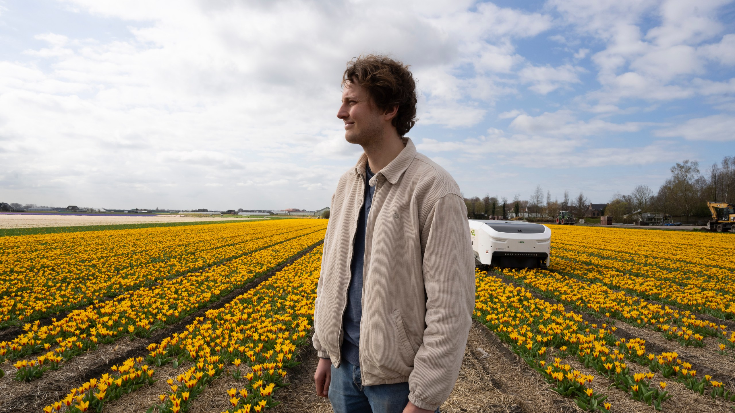 Allan Visser, a third-generation tulip farmer, is interviewed next to Theo the robot, in Noordwijkerhout, Netherlands, Tuesday, March 19, 2024. An artificial intelligence robot is a new high-tech weapon in the battle to root out disease from Dutch tulip fields as they erupt into a riot of springtime color. The robot is replacing a dwindling number of human “sickness spotters” who patrol bulb fields on the lookout for diseased flowers. (AP Photo/Peter Dejong)