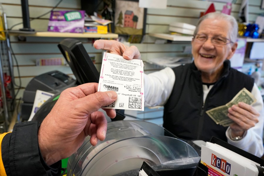 FILE - Dot Skoko, owner of Dot's Dollar More or Less shop in Mt. Lebanon, Pa., hands a customer a Mega Millions lottery ticket, Thursday, Jan. 5, 2022. The Mega Millions jackpot has reached an astounding $977 million for Friday night’s drawing after no tickets matched all six numbers drawn on Tuesday night. (AP Photo/Gene J. Puskar, File)