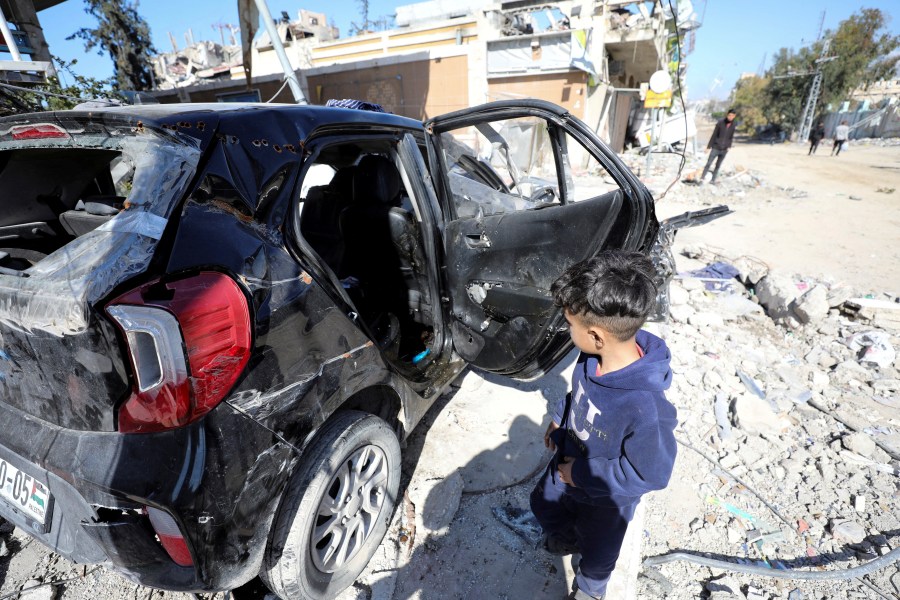 A child walks past a wrecked car full of bullet holes sitting in rubble.