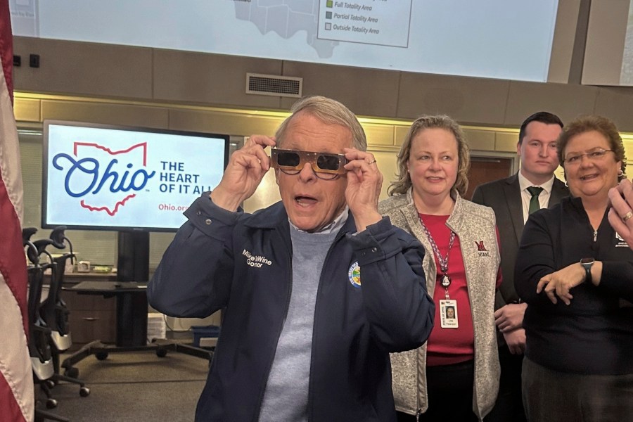 Republican Ohio Gov. Mike DeWine sports a pair of eclipse glasses while talking to reporters at the Ohio Emergency Operations Center in Columbus, Ohio, on Friday, April 5, 2024. DeWine activated the center on Friday to assist communities before, during and after Monday's total solar eclipse, which could draw hundreds of thousands of tourists to the state. (AP Photo/Julie Carr Smyth)