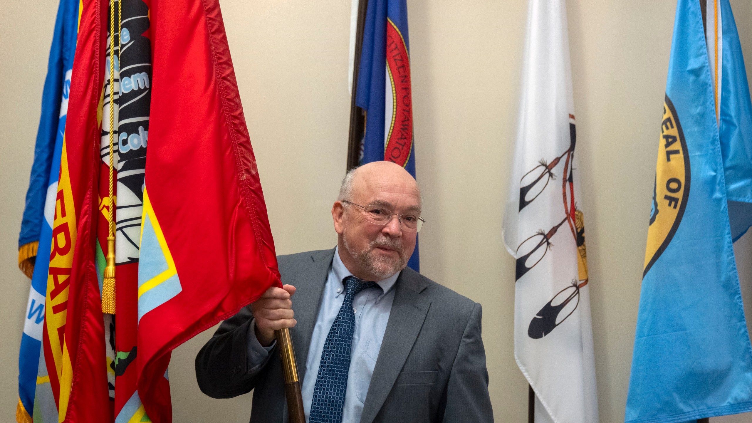 Tracy Toulou, the outgoing Director of the Office of Tribal Justice, holds the flag of the Colville Confederated Tribes, his tribal affiliation, alongside flags of tribal nations at the Department of Justice, Thursday, March 14, 2024, in Washington. For more than two decades, Toulou has confronted the serious public safety challenges facing Indian Country by working to expand the power of tribal justice systems. Today, tribal law enforcement finally has a seat at the table when federal authorities coordinate with state and local police, according to the Justice Department’s point person on Native American tribes. (AP Photo/Mark Schiefelbein)