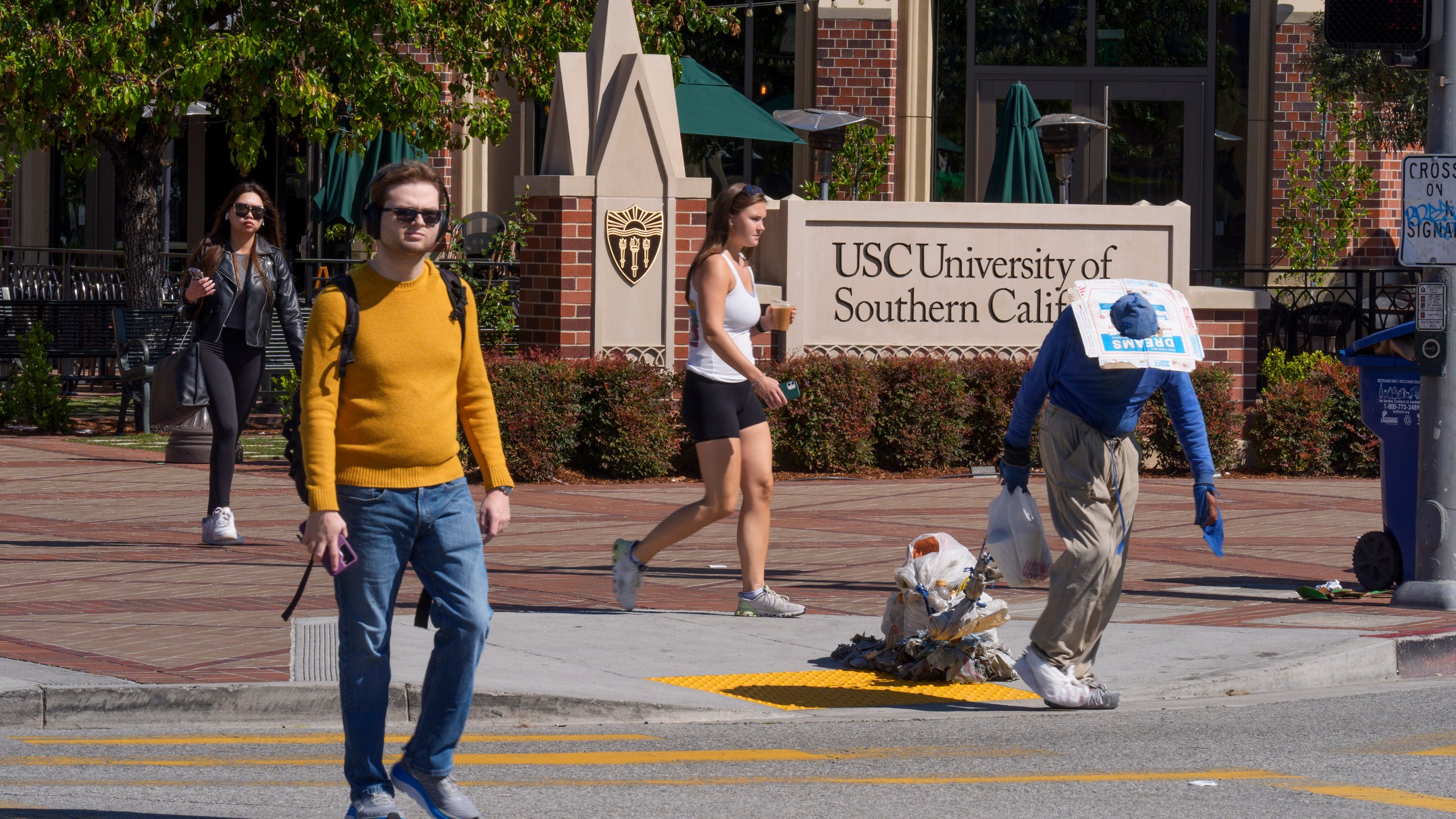 A person suffering homelessness walks past students outside the University of Southern California campus entrance in Los Angeles on Tuesday, April 16, 2024. University of Southern California officials have canceled a commencement speech by its 2024 valedictorian, a pro-Palestinian Muslim, citing "substantial risks relating to security and disruption" of the event that draws 65,000 people to campus. (AP Photo/Damian Dovarganes)