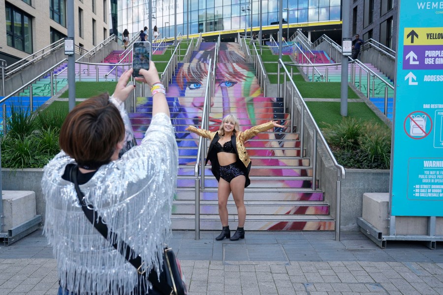 A fan poses for a photo by a staircase with the image of Taylor Swift, outside Wembley Stadium ahead of her first London concert, during the Eras Tour, in London, Friday June 21, 2024. (Lucy North/PA via AP)