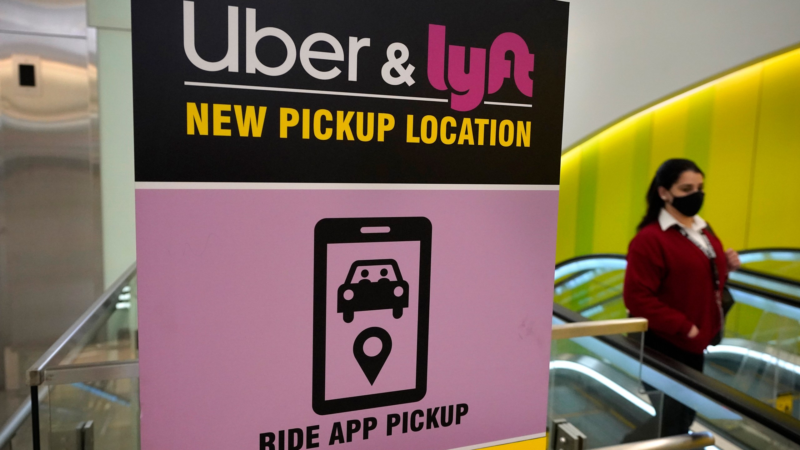 FILE - A passerby walks past a sign offering directions to an Uber and Lyft ride pickup location at an airport, Feb. 9, 2021, in Boston. Drivers for Uber and Lyft will earn a minimum pay standard of $32.50 per hour under a settlement announced, Thursday, June 27, 2024, by Massachusetts Attorney General Andrea Campbell, a deal that also includes a suite of benefits and protections. (AP Photo/Steven Senne, File)