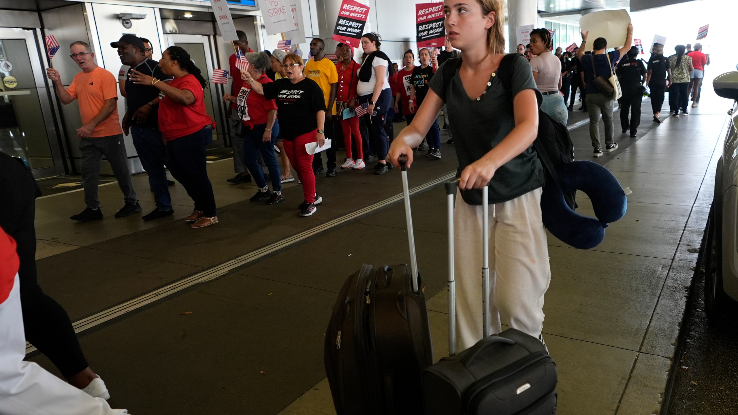 A traveler walks through the departure area as airline catering workers who are employed by Gate Gourmet picket with their supporters, calling for a new union contract with raises and affordable health insurance, Wednesday, July 3, 2024, at Miami International Airport in Miami. A long Fourth of July holiday weekend is expected to create new travel records. (AP Photo/Lynne Sladky)