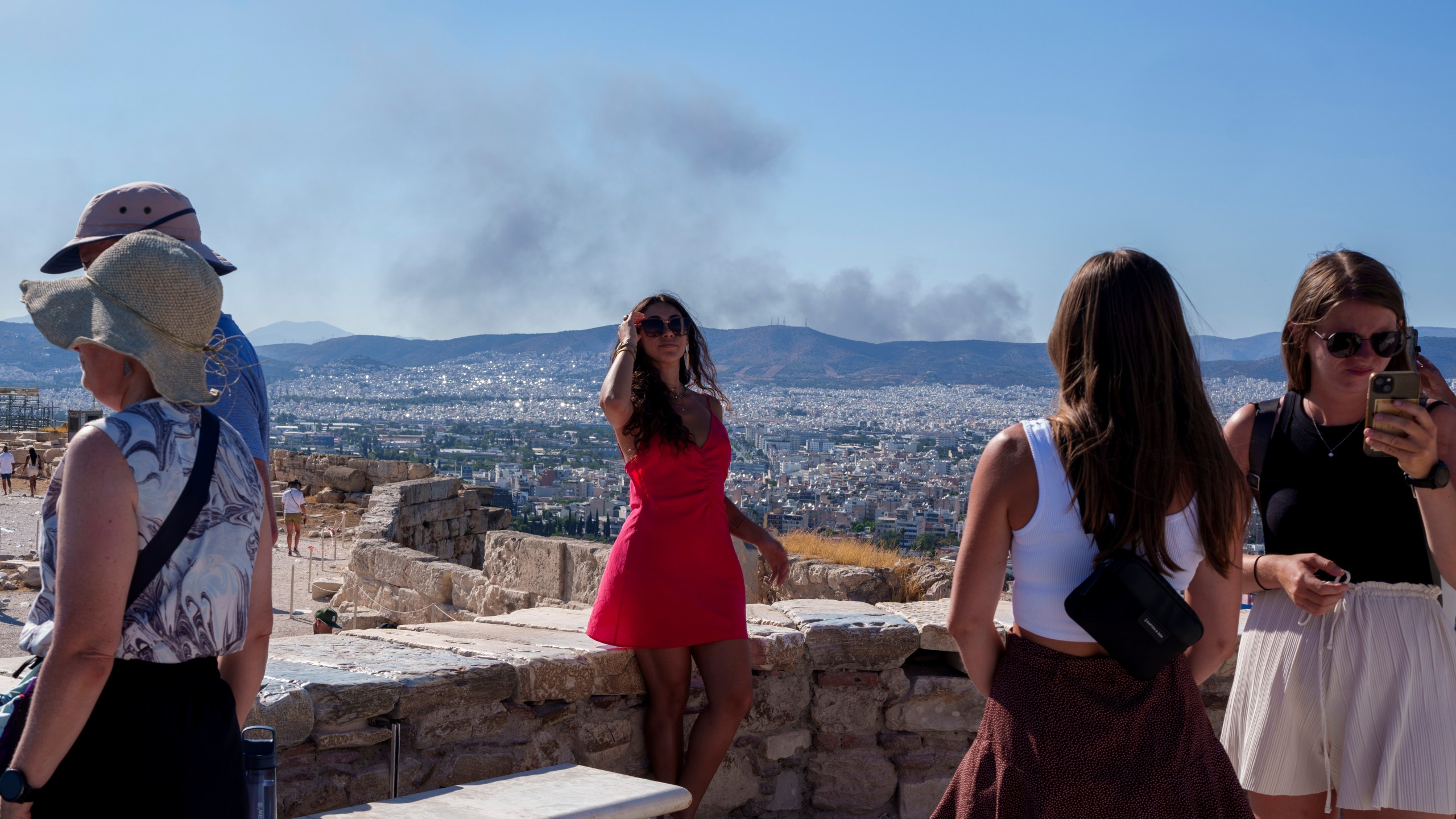 A woman poses for a photo with smoke from a fire at the background, during a hot, windy day at Acropolis hill, in Athens, Tuesday, July 16, 2024. Much of Greece was also sweltering in a heat wave due to last until the end of the week, with temperatures in some areas forecast to reach 42 degrees Celsius (over 107 degrees Fahrenheit).(AP Photo/Petros Giannakouris)