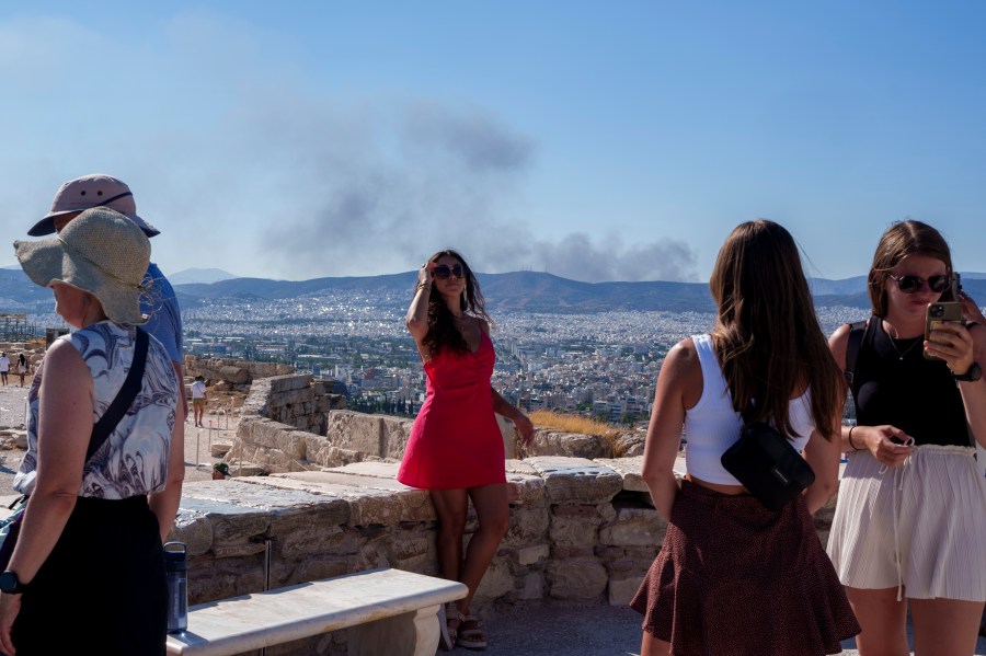 A woman poses for a photo with smoke from a fire at the background, during a hot, windy day at Acropolis hill, in Athens, Tuesday, July 16, 2024. Much of Greece was also sweltering in a heat wave due to last until the end of the week, with temperatures in some areas forecast to reach 42 degrees Celsius (over 107 degrees Fahrenheit).(AP Photo/Petros Giannakouris)