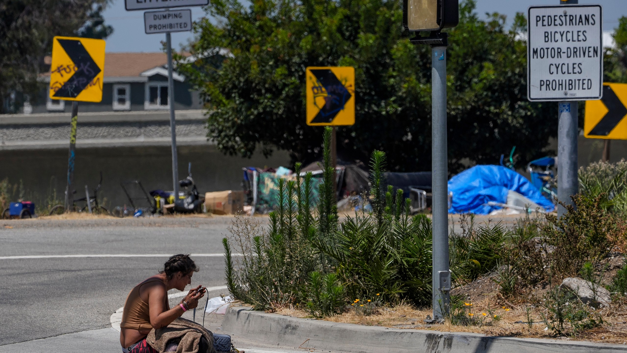 A homeless woman sits on the street at the entrance to an off-ramp to the CA-105 freeway next to a homeless encampment Friday, July 26, 2024, in Los Angeles. (AP Photo/Damian Dovarganes)