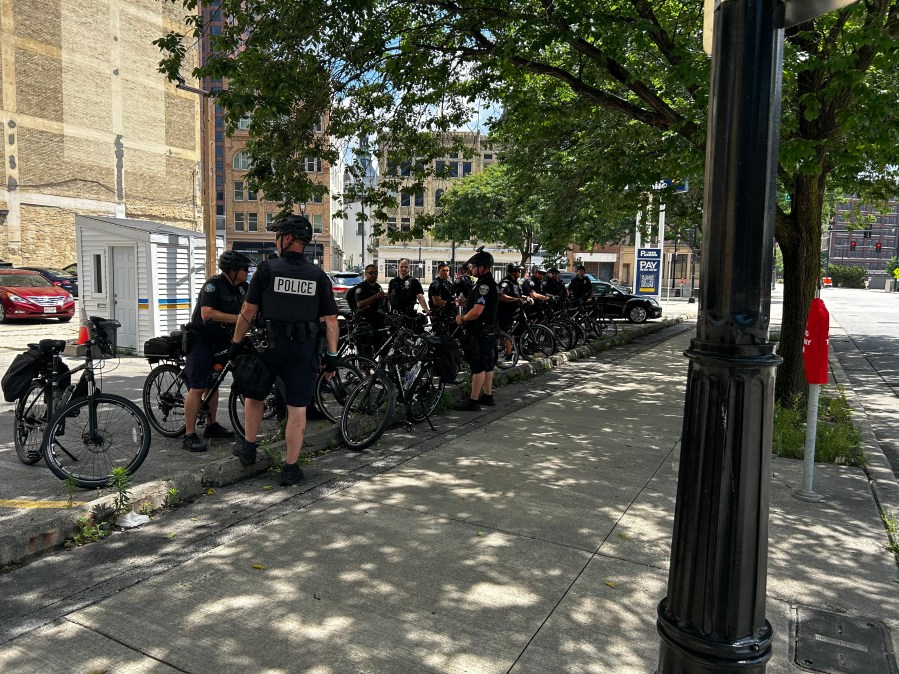 Milwaukee police officers on bicycles wear bike helmets and black shirts with "POLICE" on the back as they stand outside near the RNC secure zone.