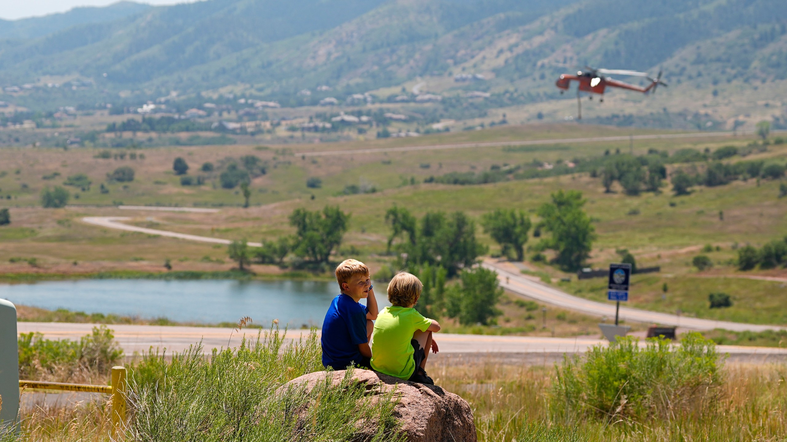 Young boys sit on a rock to watch as a helicopter lands on a nearby lake to collect water to drop on ridges as the Quarry wildfire burns in the foothills near the Ken Caryl Ranch development Thursday, Aug. 1, 2024, southwest of Littleton, Colo. (AP Photo/David Zalubowski)