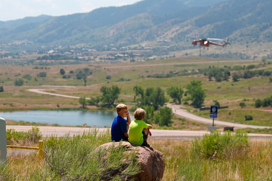 Young boys sit on a rock to watch as a helicopter lands on a nearby lake to collect water to drop on ridges as the Quarry wildfire burns in the foothills near the Ken Caryl Ranch development Thursday, Aug. 1, 2024, southwest of Littleton, Colo. (AP Photo/David Zalubowski)