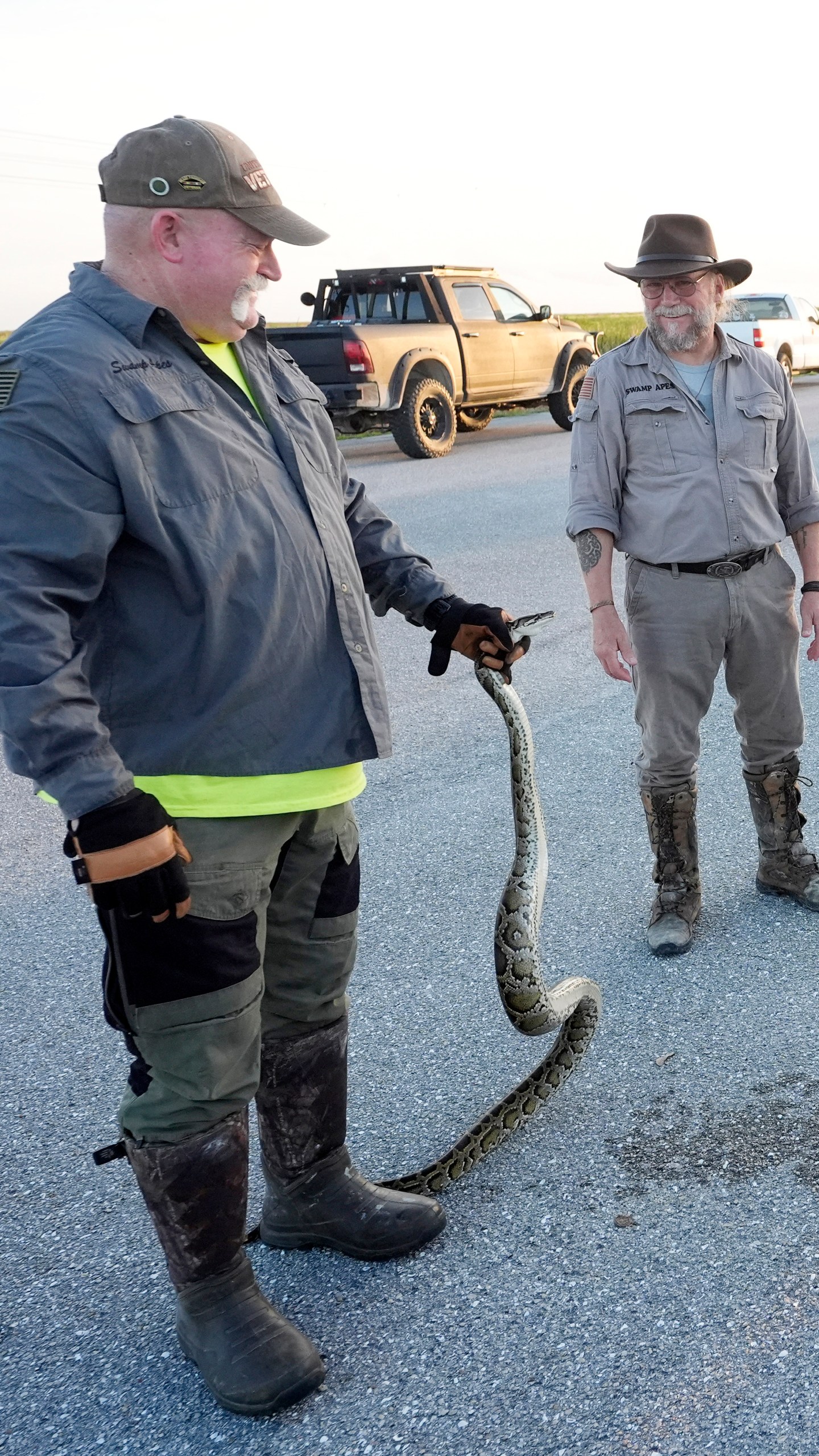 Contractors with the Florida Fish and Wildlife Conservation Commission, Thomas Aycock, left, and Tom Rahill, founder of the Swamp Apes, a veterans therapy nonprofit, show off an invasive Burmese python caught earlier, as they wait for sunset to hunt pythons, Tuesday, Aug. 13, 2024, in the Florida Everglades. (AP Photo/Wilfredo Lee)