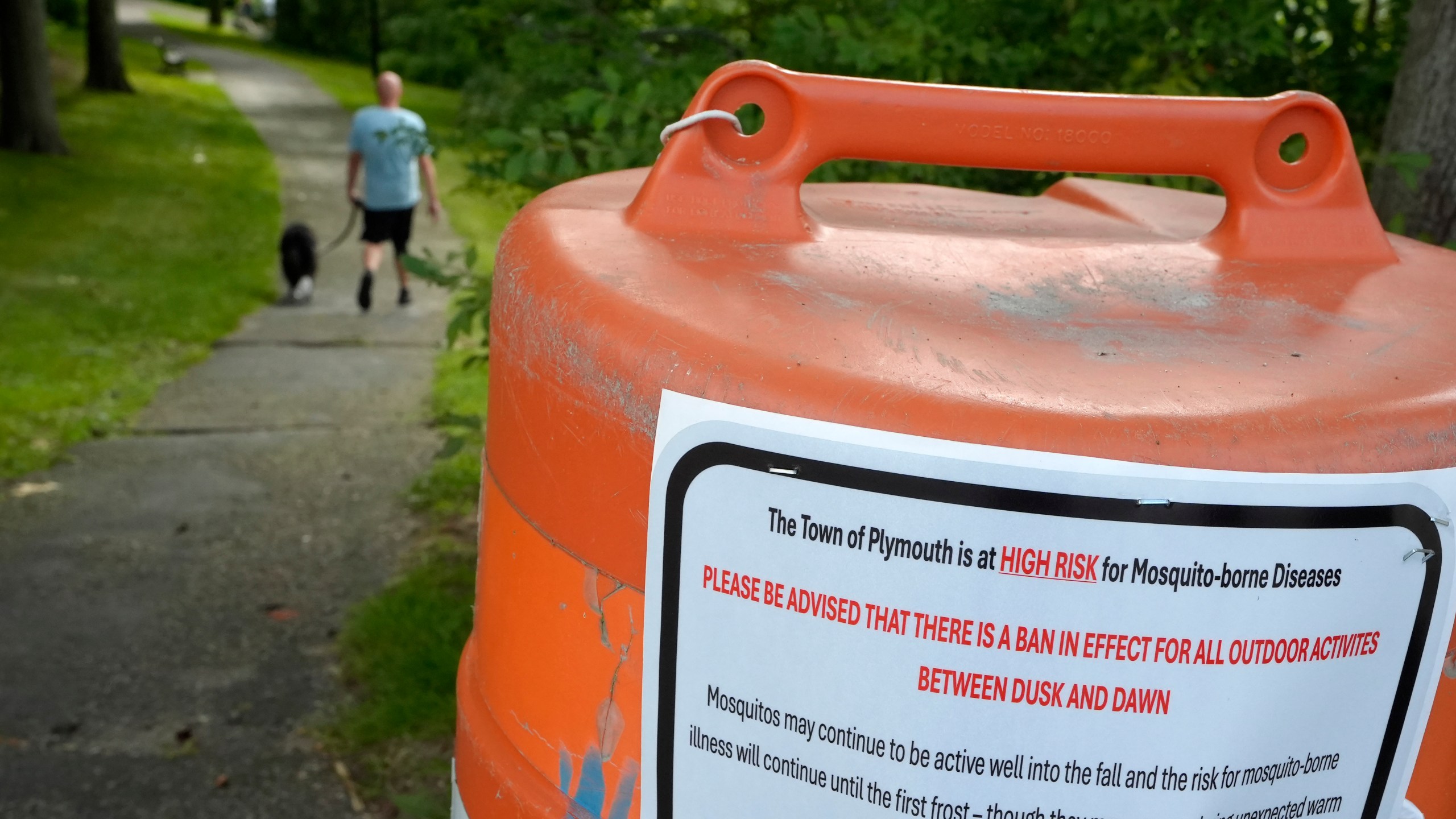 A passer-by walks a dog, Monday, Aug. 26, 2024, along a walkway, in Plymouth, Mass., near a sign that advises people of a ban in effect for outdoor activity between dusk and dawn due to the risk of exposure to mosquito-borne diseases. (AP Photo/Steven Senne)