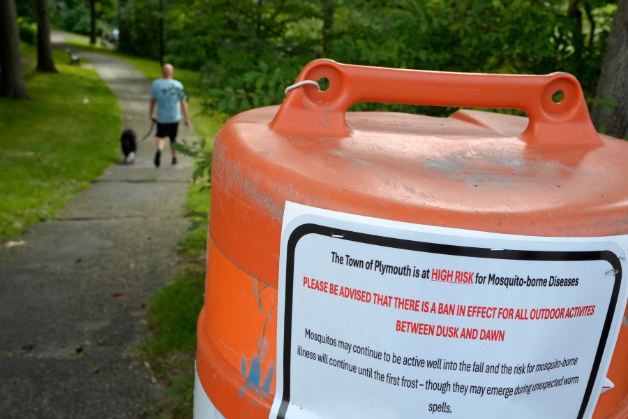 A passer-by walks a dog, Monday, Aug. 26, 2024, along a walkway, in Plymouth, Mass., near a sign that advises people of a ban in effect for outdoor activity between dusk and dawn due to the risk of exposure to mosquito-borne diseases. (AP Photo/Steven Senne)