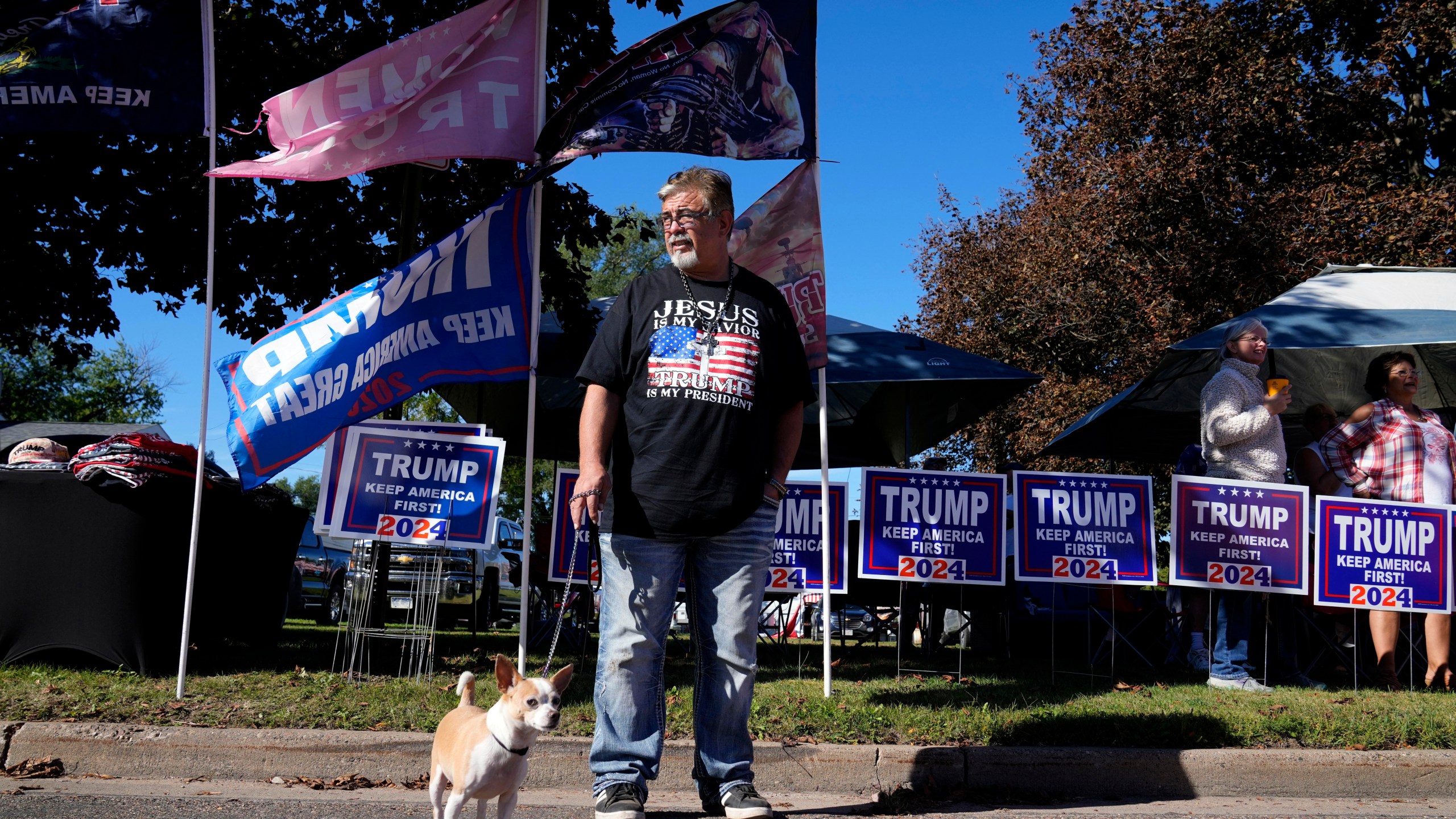 Merchandise vendor Sam Smith, of Sarasota, Fla., stands with his dog Milo while waiting for Republican presidential nominee former President Donald Trump to arrive at a rally, Saturday, Sept. 28, 2024, in Prairie du Chien, Wis. (AP Photo/Charlie Neibergall)