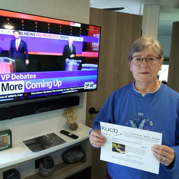 Mary Nelson, of Mead, Wash., poses for a photo at her home on Wednesday, Oct. 2, 2024, as a local TV station broadcasts a news story about the vice presidential debate. Nelson is holding a printout of a radio station website news story that says she is thought to be the last person in the U.S. to cast a vote in the 2012 presidential election when she voted for Mitt Romney just before the polls closed in the remote village of Adak, Alaska. (AP Photo/Ted S. Warren)