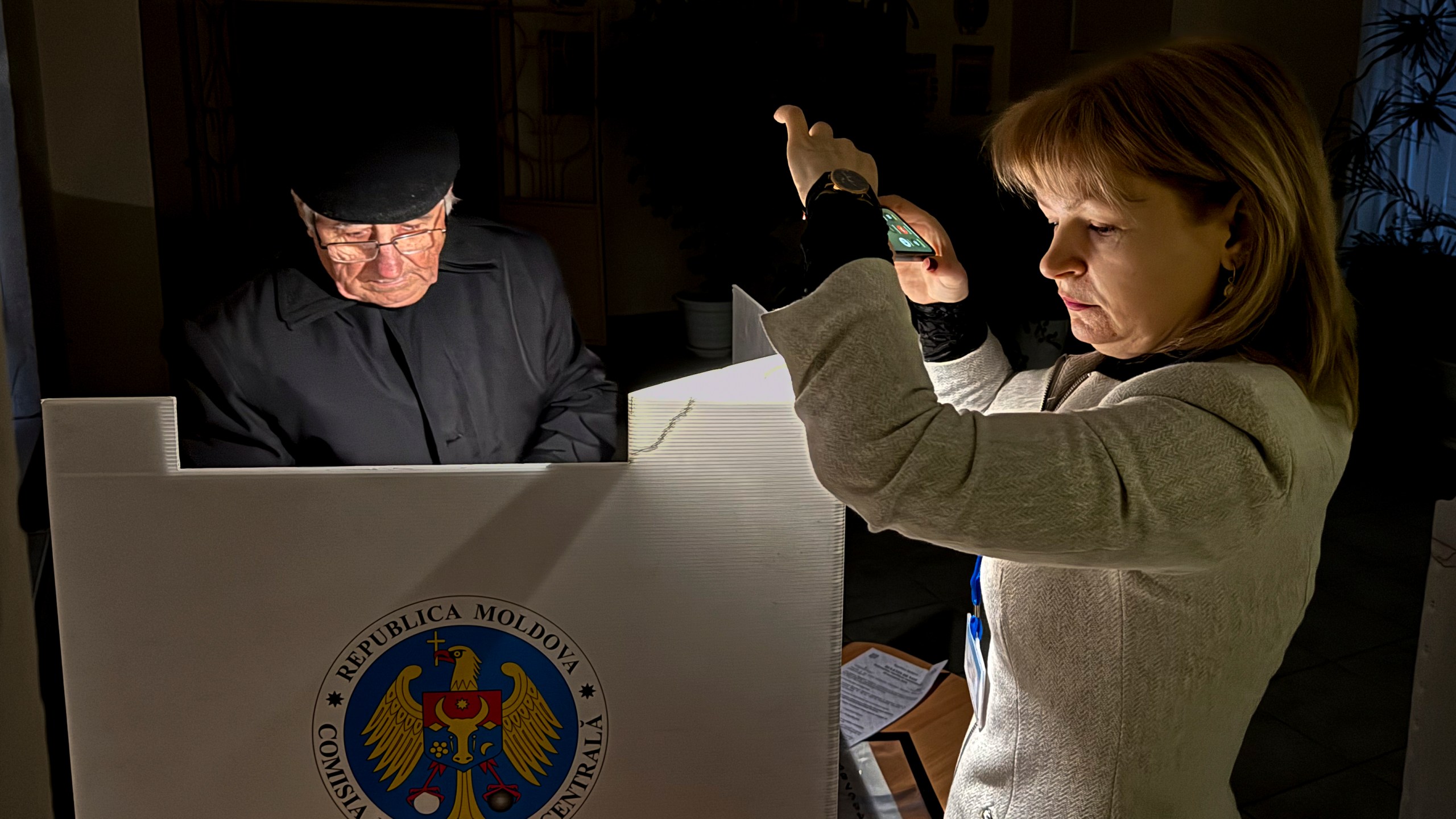 An election official assists a voter with her mobile phone flashlight during a power failure in Chisinau, Moldova, Sunday, Oct. 20, 2024, during a presidential election and a referendum on whether to enshrine in the Constitution the country's path to European Union membership. (AP Photo/Nicolae Dumitrache)