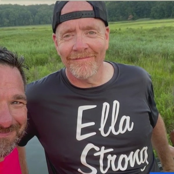 Mike Downey wears a shirt that says "Ella Strong" as he stands between two people, all smiling for a group photo.