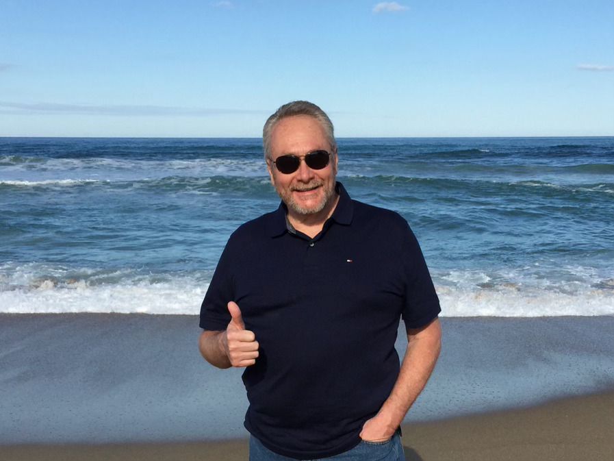 An older man on a beach giving a thumbs up.