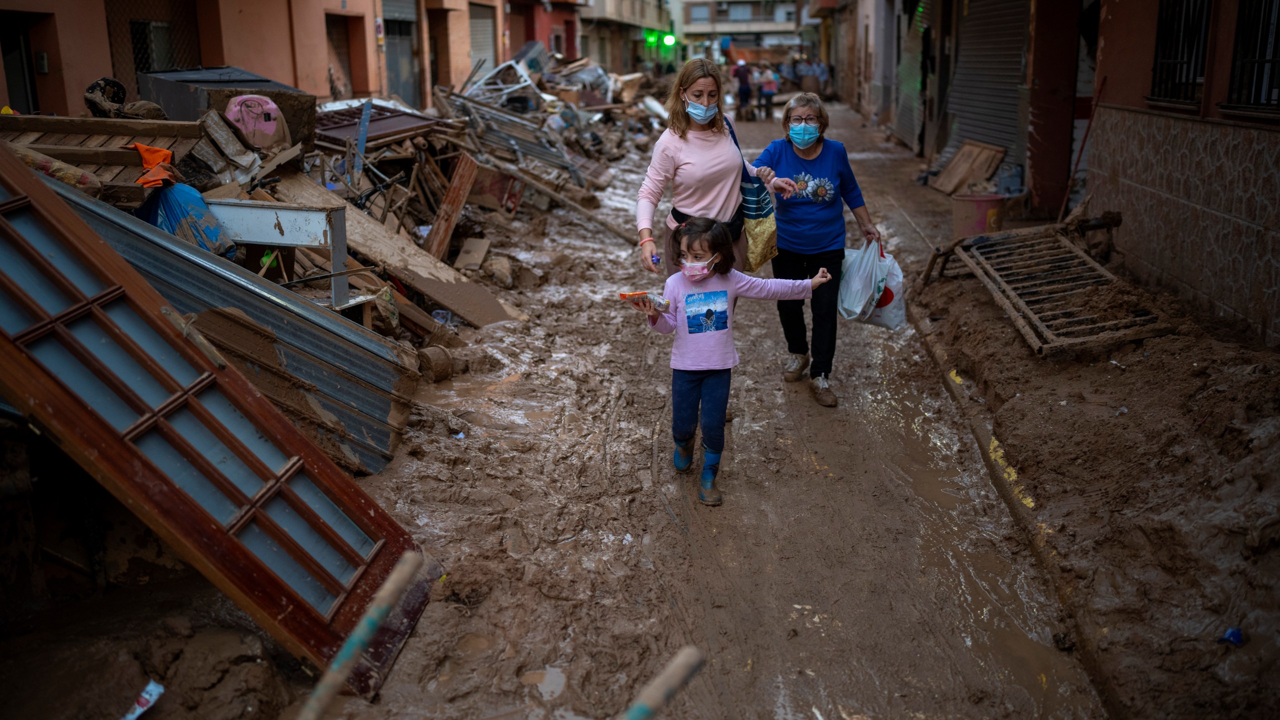 A child walks with her mother and grandmother through a street with piled furniture and rubbish on the sides, in an area affected by floods in Paiporta, Valencia, Spain, Tuesday, Nov. 5, 2024. (AP Photo/Emilio Morenatti)