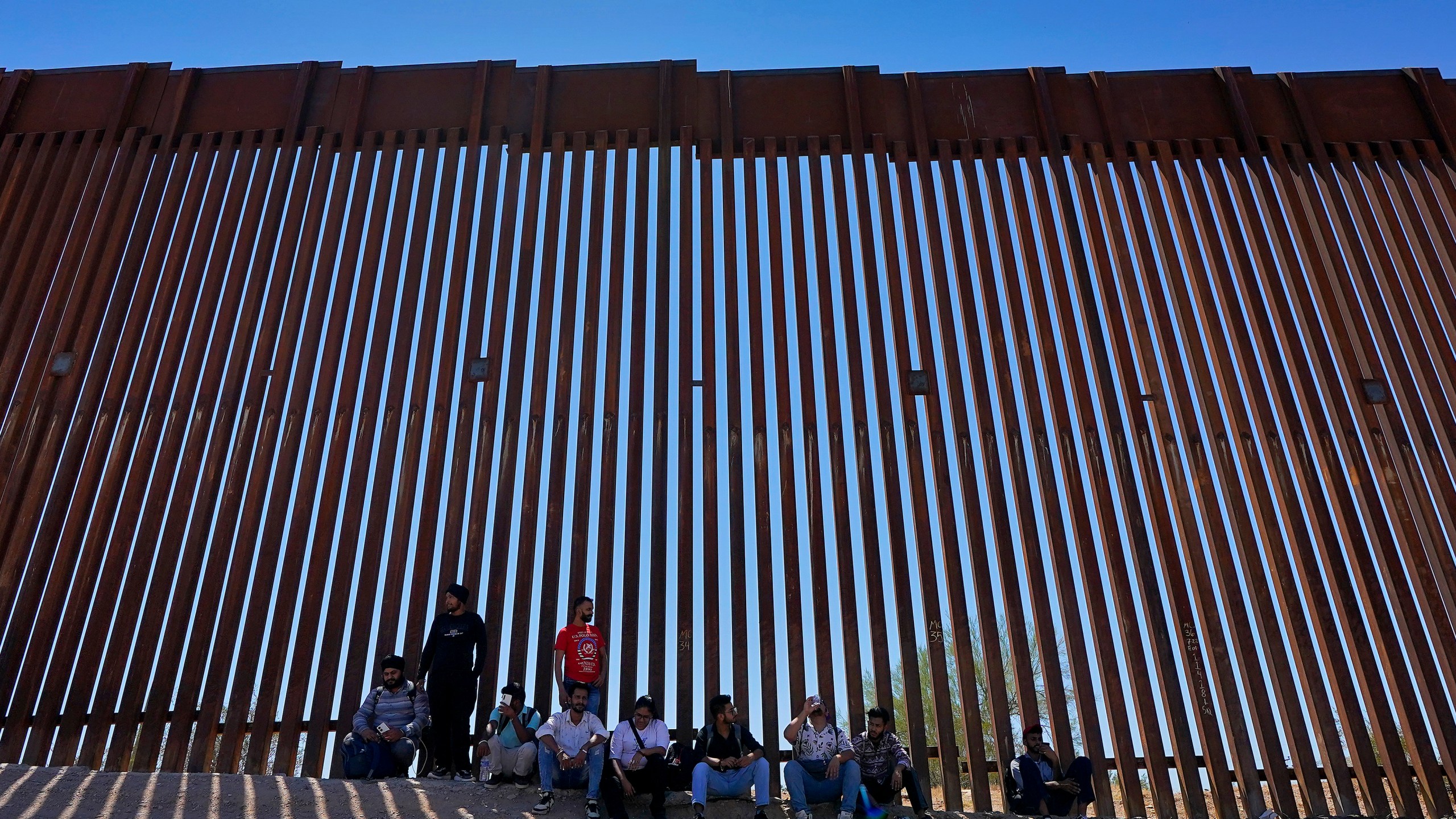 FILE - A group claiming to be from India sit in the shade of the border wall as they wait to be picked up by Border Patrol after crossing through the border fence in the Tucson Sector of the U.S.-Mexico border, Aug. 29, 2023, in Organ Pipe Cactus National Monument near Lukeville, Ariz. (AP Photo/Matt York, File)