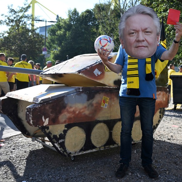 FILE - A dummy tank stands next to an activist wearing a mask with the likeness of Rheinmetall CEO Armin Papperger and holding up a red card ahead of the soccer game between Borussia Dortmund and Eintracht Frankfurt, in Dortmund, Germany, Saturday Aug. 24, 2024. (Bernd Thissen/dpa via AP, File)