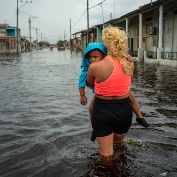 FILE - A woman carries a child as she wades through a street flooded in the passing of Hurricane Helene, in Batabano, Mayabeque province, Cuba, Sept. 26, 2024. (AP Photo/Ramon Espinosa, File)