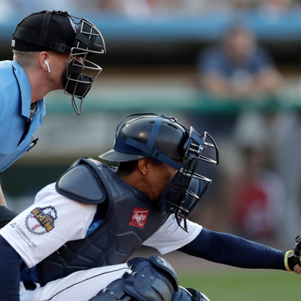 FILE -Home plate umpire Brian deBrauwere, left, huddles behind Freedom Division catcher James Skelton, of the York Revolution, as the official wears an earpiece during the first inning of the Atlantic League All-Star minor league baseball game, Wednesday, July 10, 2019, in York, Pa. Major League Baseball will test robot umpires as part of a challenge system during spring training at 13 ballparks hosting 19 teams, which could lead to regular-season use in 2026. (AP Photo/Julio Cortez, File)