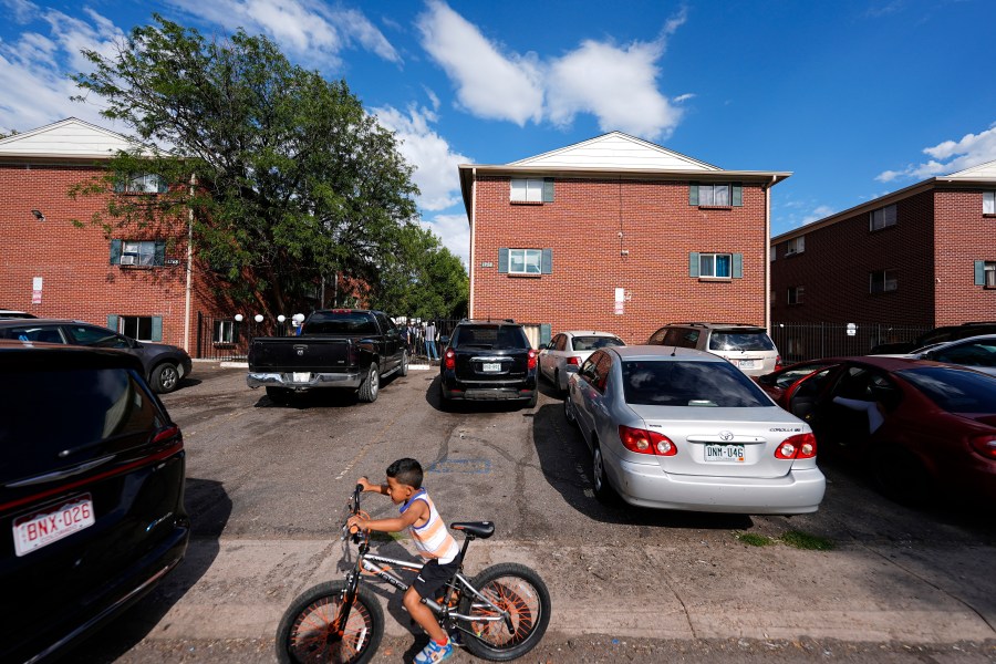 FILE - A boy rides his bicycle past apartment buildings as a rally staged by the East Colfax Community Collective is held in the courtyard to address chronic problems in the apartment buildings occupied by people displaced from their home countries in central and South America, Sept. 3, 2024, in Aurora, Colo. (AP Photo/David Zalubowski, File)