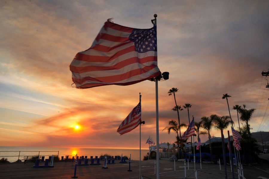 FILE - Flags fly under heavy winds before sunset as a plume of smoke from the Franklin Fire rises over the ocean Tuesday, Dec. 10, 2024, in Malibu, Calif. (AP Photo/Damian Dovarganes,File)