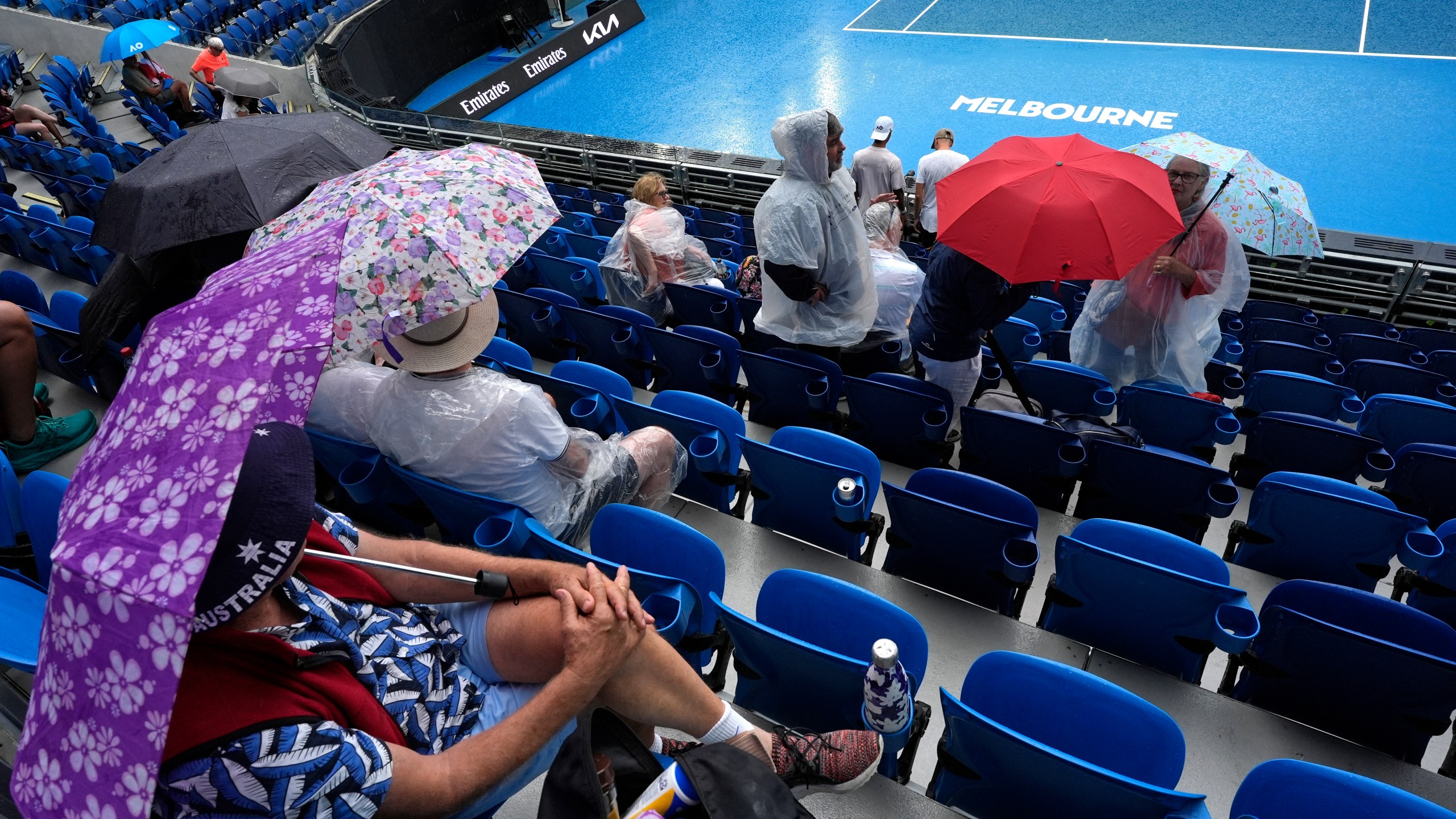 Spectators wait under umbrellas as rain suspends play during first round matches at the Australian Open tennis championship in Melbourne, Australia, Sunday, Jan. 12, 2025. (AP Photo/Asanka Brendon Ratnayake)