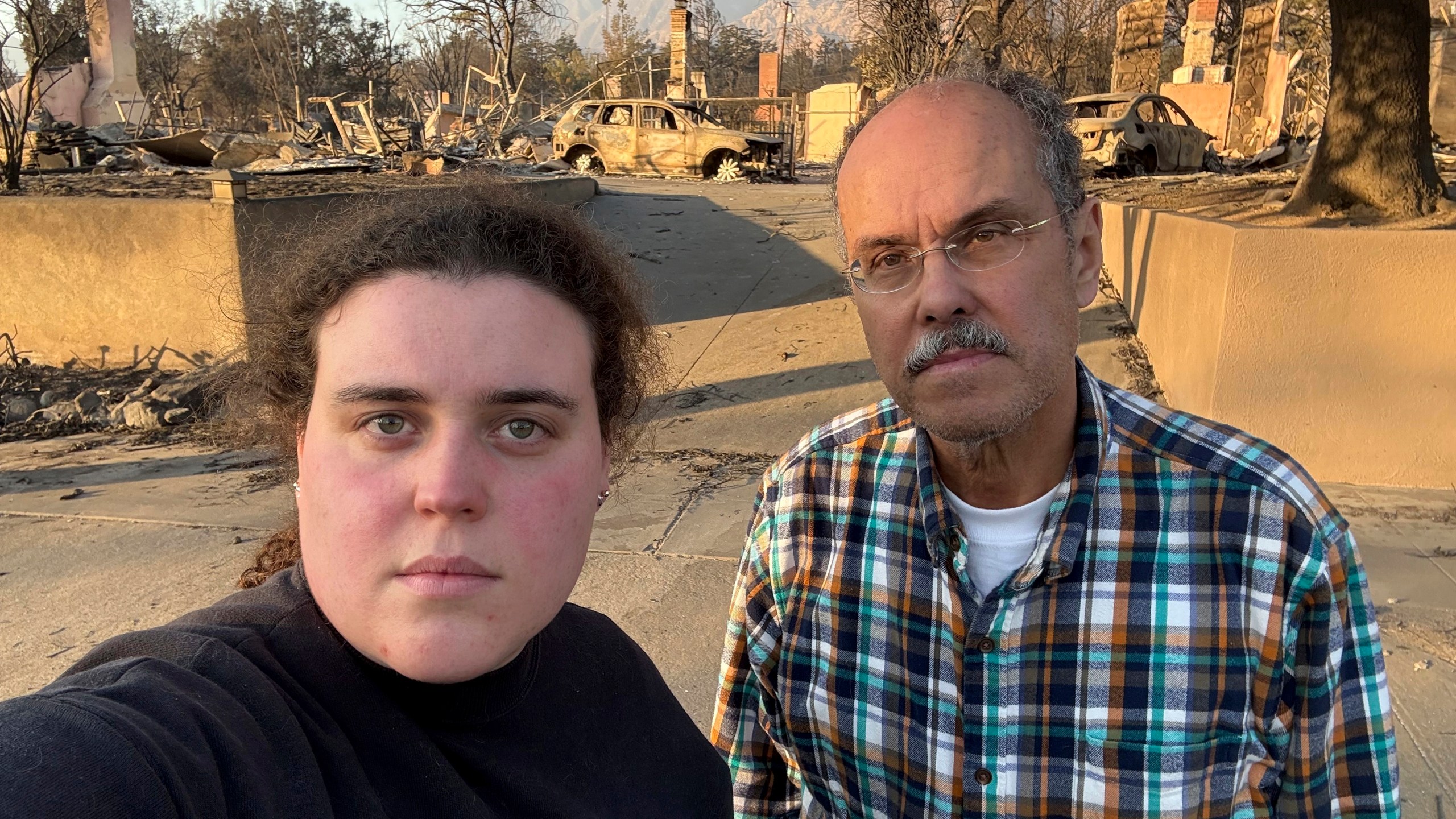 Vanessa Prata and her father, Aluizio Prata, pose for a self-portrait in Altadena, Calif., on Saturday, Jan. 11, 2025, with damage from the Eaton fire behind them. (Vanessa Prata via AP)