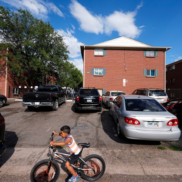 FILE - A boy rides his bicycle past apartment buildings as a rally staged by the East Colfax Community Collective is held in the courtyard to address chronic problems in the apartment buildings occupied by people displaced from their home countries in central and South America, Sept. 3, 2024, in Aurora, Colo. (AP Photo/David Zalubowski, File)